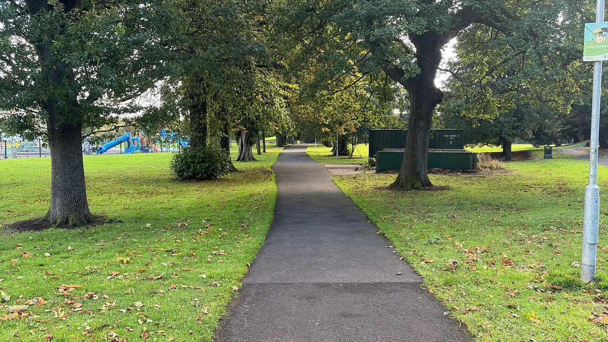 A tree-lined path through a public park, with a play area beyond